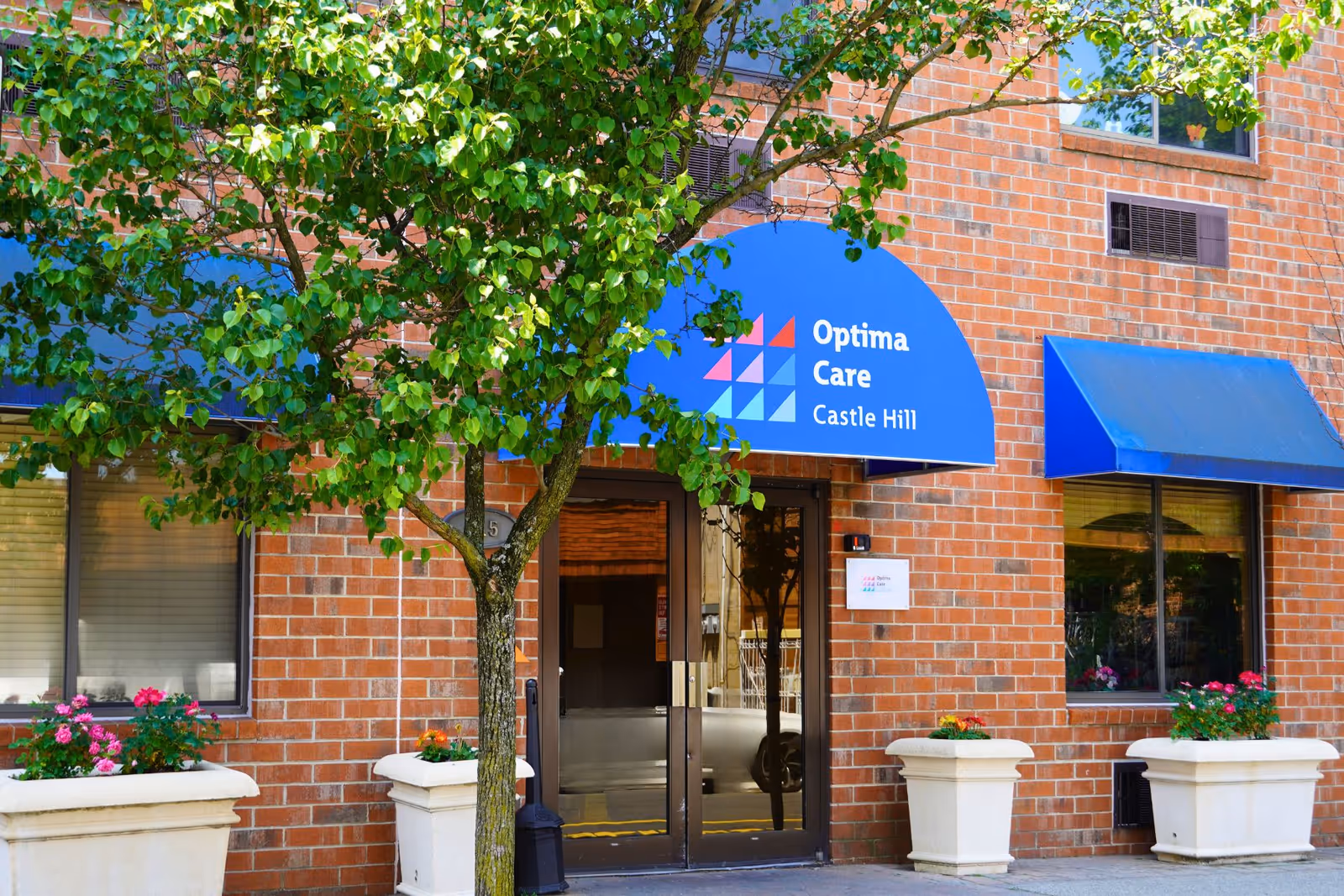 Brick building entrance with a blue awning reading "Optima Care Castle Hill", potted flowers, and a tree in front.