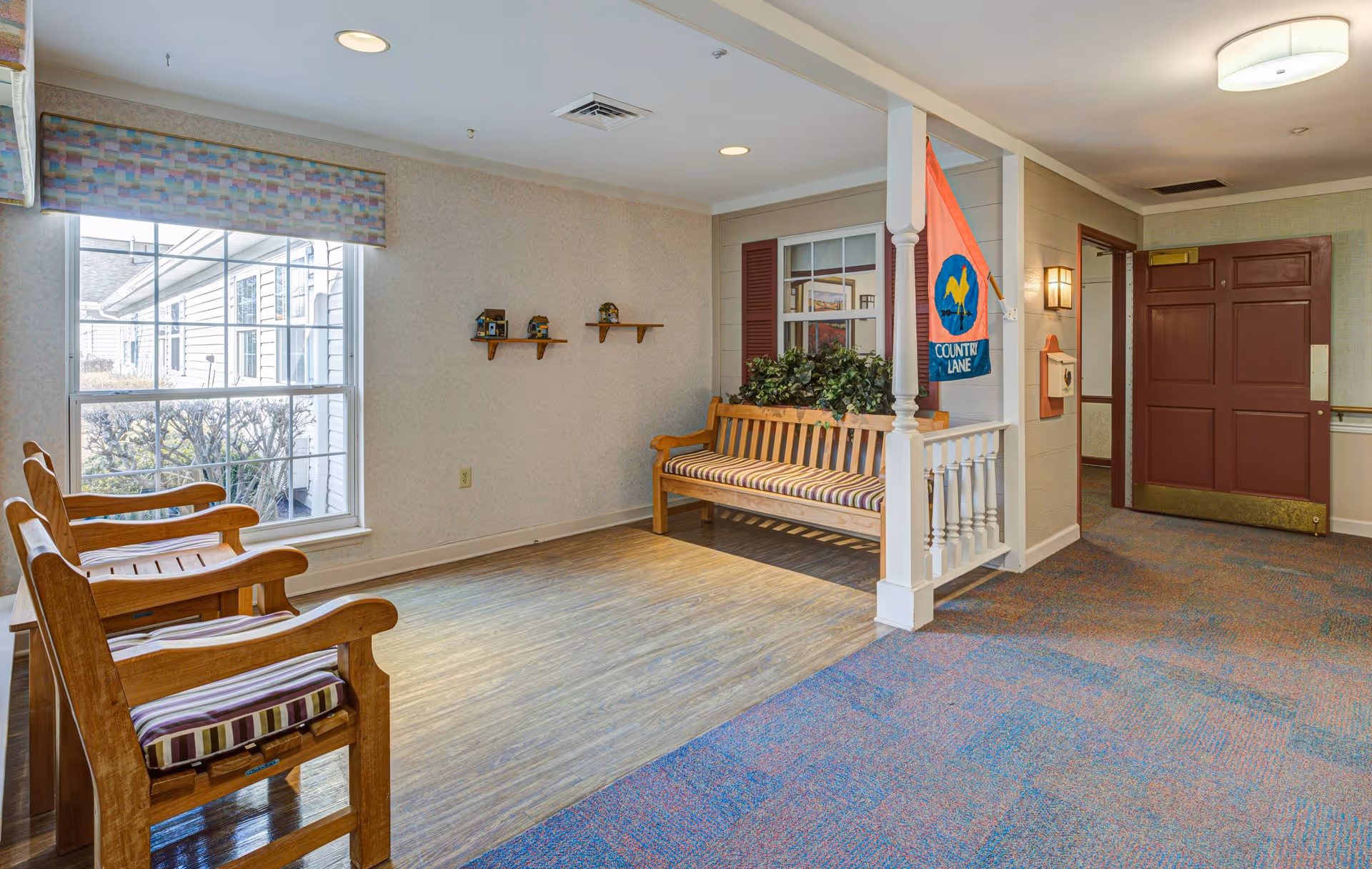 Interior view of a senior living facility common area with wooden chairs and a wooden bench with striped cushions. There is a large window with a patterned valance, small decorative shelves on the wall, a flag with the text 'COUNTRY LANE,' and a maroon door in the background. The floor has a combination of wood and carpeted sections.
