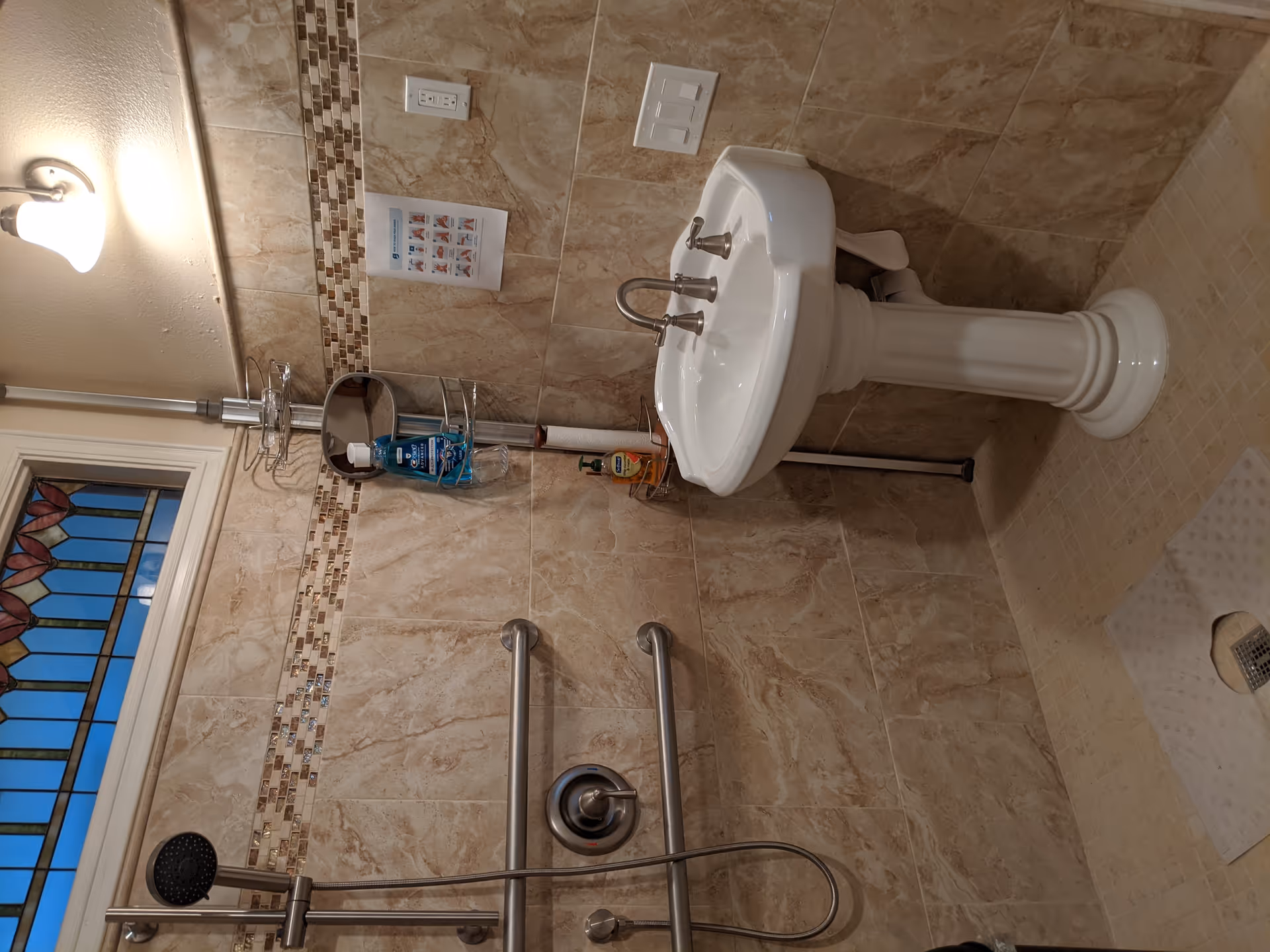 A bathroom with beige marble tiles on the walls and floor, featuring a white pedestal sink with silver faucets, a shower area with a handheld showerhead, two horizontal grab bars, and a small corner shelf holding toiletries. There is a stained glass window above the shower and a light fixture on the wall above the sink. A paper towel roll and a hand sanitizer bottle are also visible on the shelf.