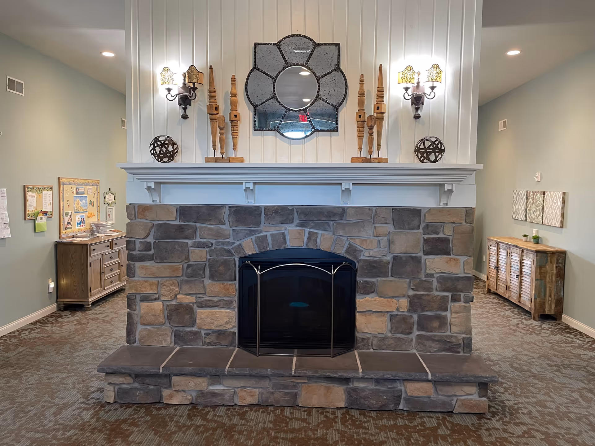Interior view of a cozy common area featuring a stone fireplace with a white wooden mantle. Above the mantle is a decorative mirror with a unique geometric design, flanked by two wall sconces with lampshades. The mantle is decorated with wooden spindle ornaments and spherical decor pieces. On either side of the fireplace, there are wooden cabinets against light green walls, with various decorations and bulletin boards.