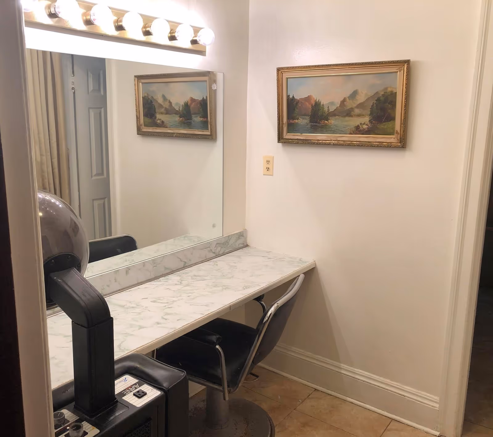 A small vanity area with a large mirror, a marble-patterned countertop, and a black salon chair. Above the mirror are five round light bulbs. On the wall next to the mirror is a framed landscape painting of mountains and trees. The floor is tiled and the walls are painted white.