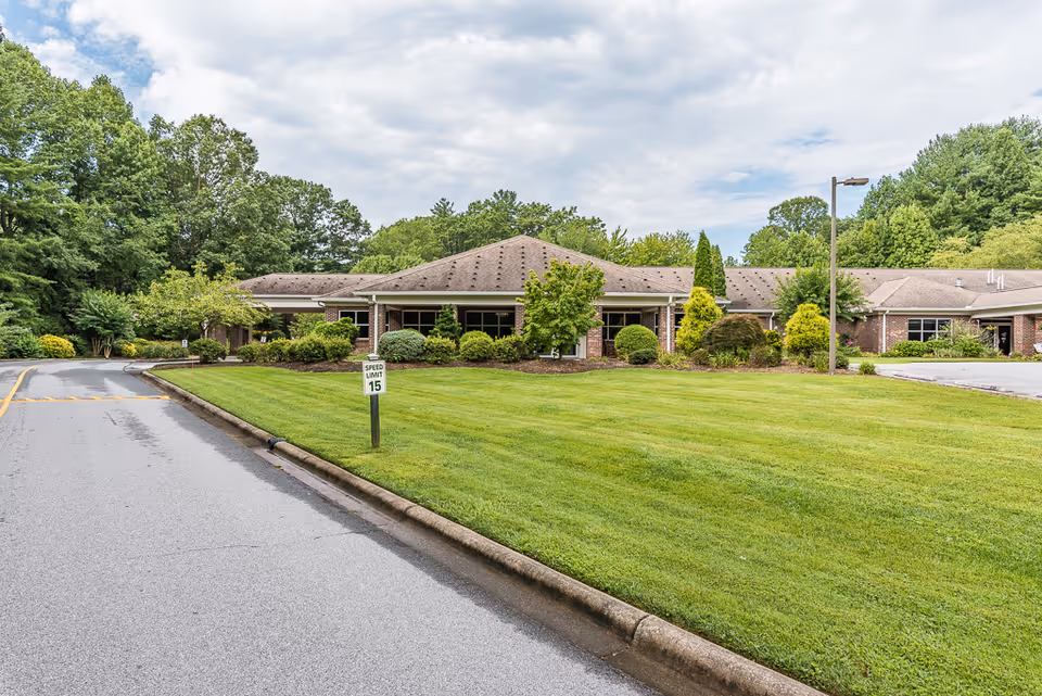 Exterior view of a single-story brick building surrounded by well-maintained green lawns and various trees and shrubs under a partly cloudy sky. A paved road with a speed limit sign of 15 mph runs alongside the building.