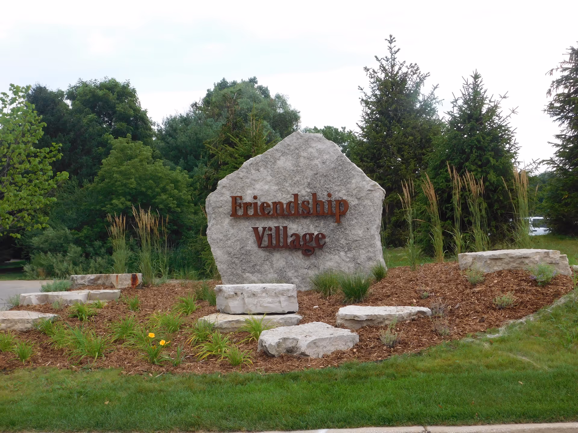 A large stone monument with the words 'Friendship Village' mounted on it, surrounded by landscaped greenery, plants, and trees in the background.