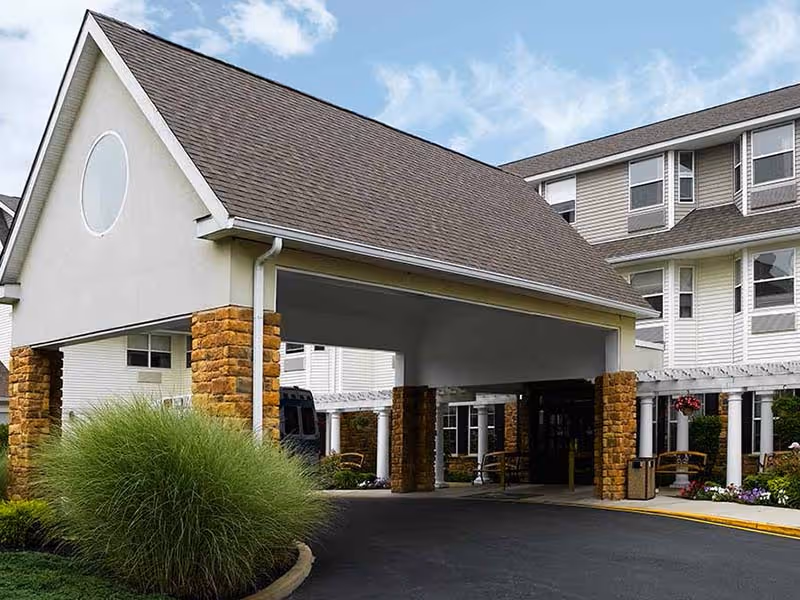 Entrance to a senior living facility with a covered drop-off area supported by stone pillars, surrounded by landscaped greenery and a multi-story building in the background under a partly cloudy sky.