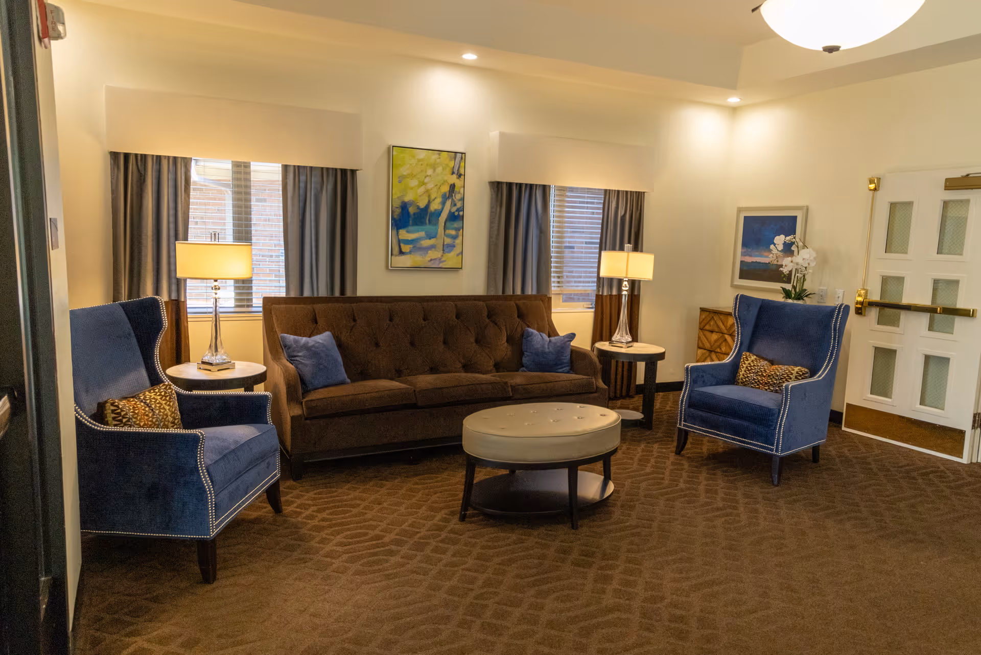 A cozy living room with a brown tufted sofa adorned with blue pillows, two blue upholstered armchairs with patterned cushions, two round side tables each holding a lamp, a round ottoman coffee table, two windows with curtains, and framed artwork on the walls. The room has a brown patterned carpet and a white ceiling light fixture.