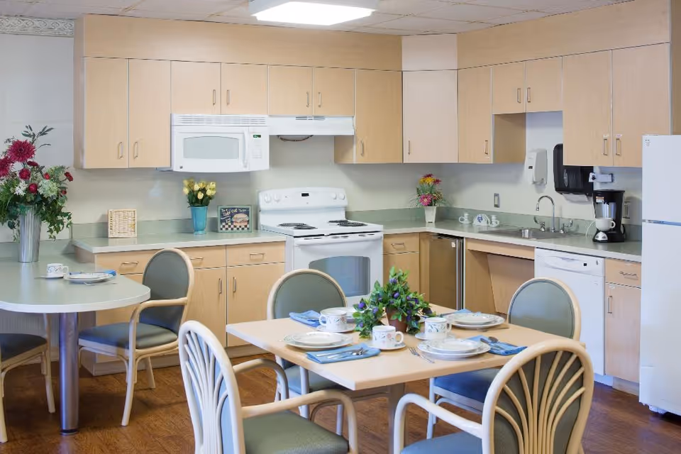 A well-lit kitchen area with light wood cabinets, a white stove, microwave, dishwasher, and refrigerator. There are two tables with chairs around them, set with plates, cups, and napkins. Decorative flowers are placed on the tables and countertops.