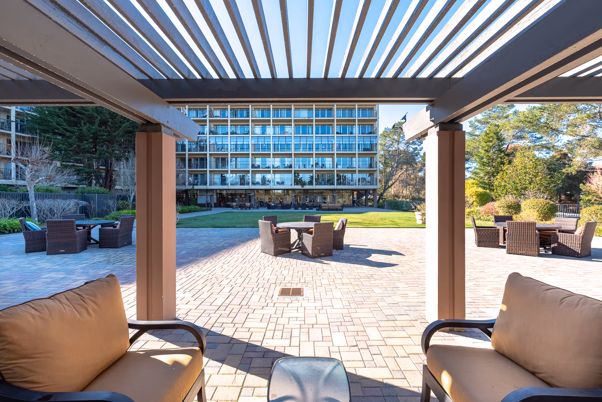 Outdoor patio area with cushioned chairs and tables under a pergola, overlooking a lawn and a multi-story building with balconies in the background. Trees and shrubs surround the patio area.