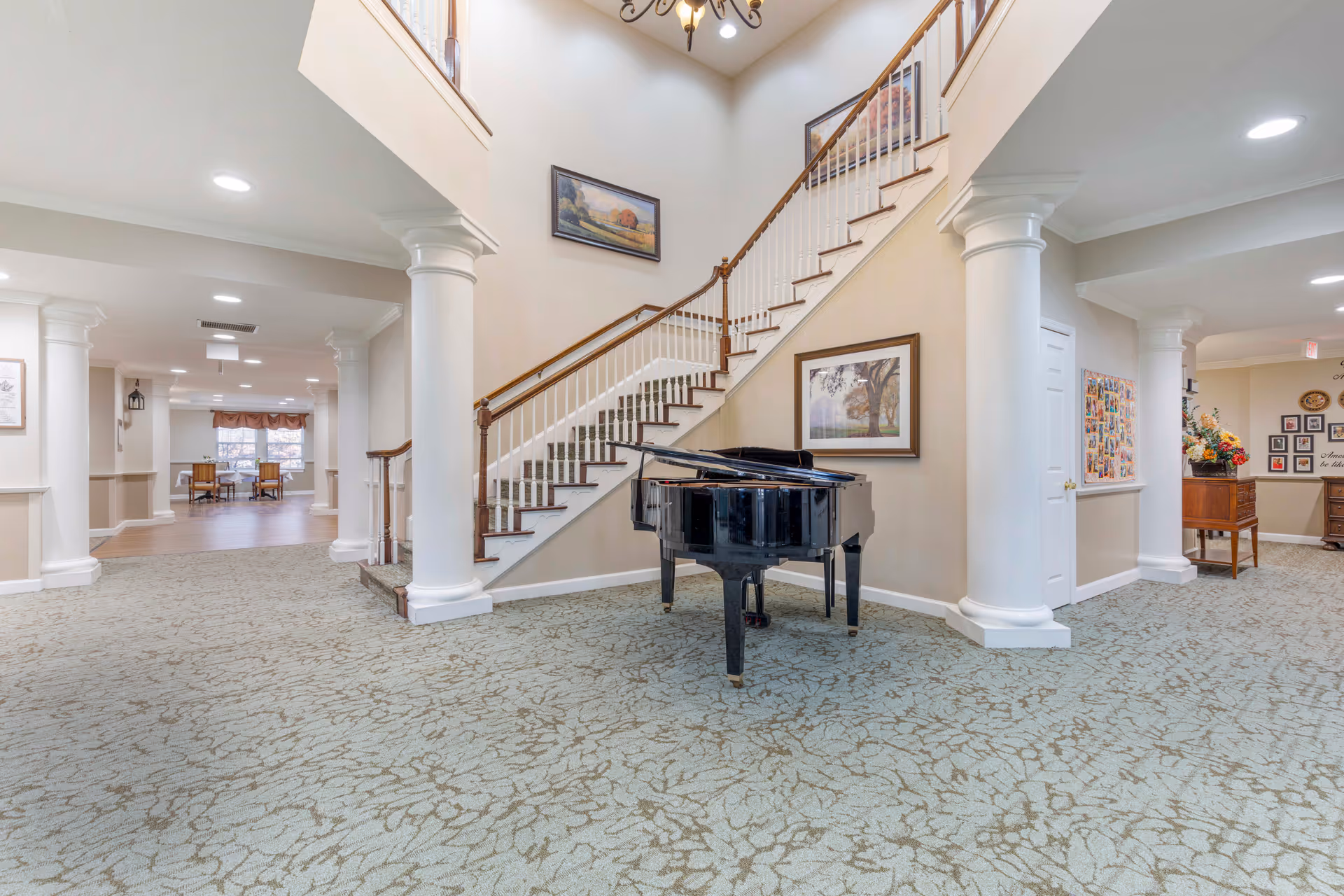 Interior view of a senior living facility lobby area featuring a grand piano in front of a staircase with wooden handrails and white balusters. The space has beige walls, decorative columns, framed artwork, and patterned carpet flooring. In the background, there is a seating area with tables and chairs near large windows.