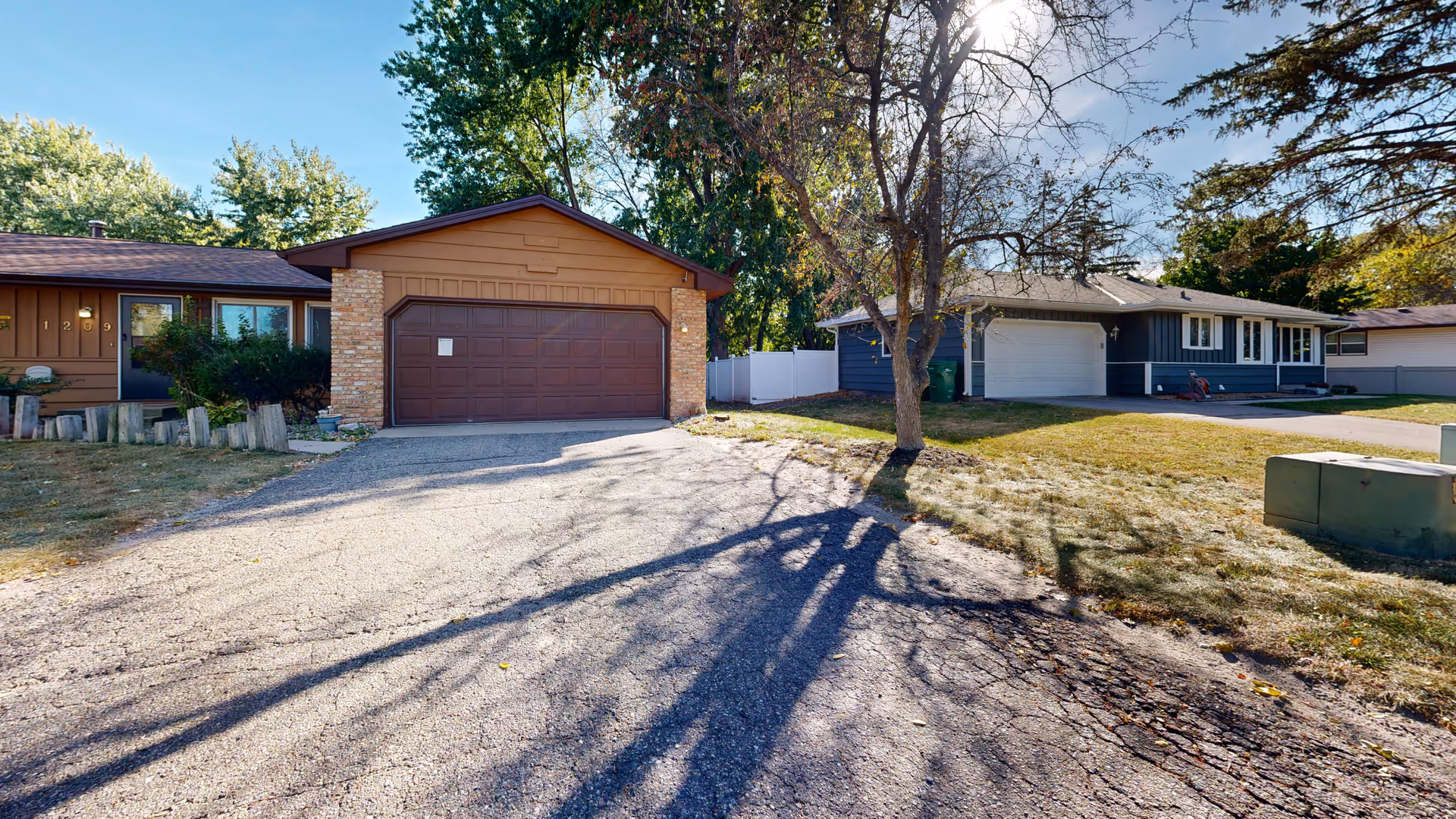 Two single-story homes with garages on a residential street, a tree casting long shadows across the driveway.