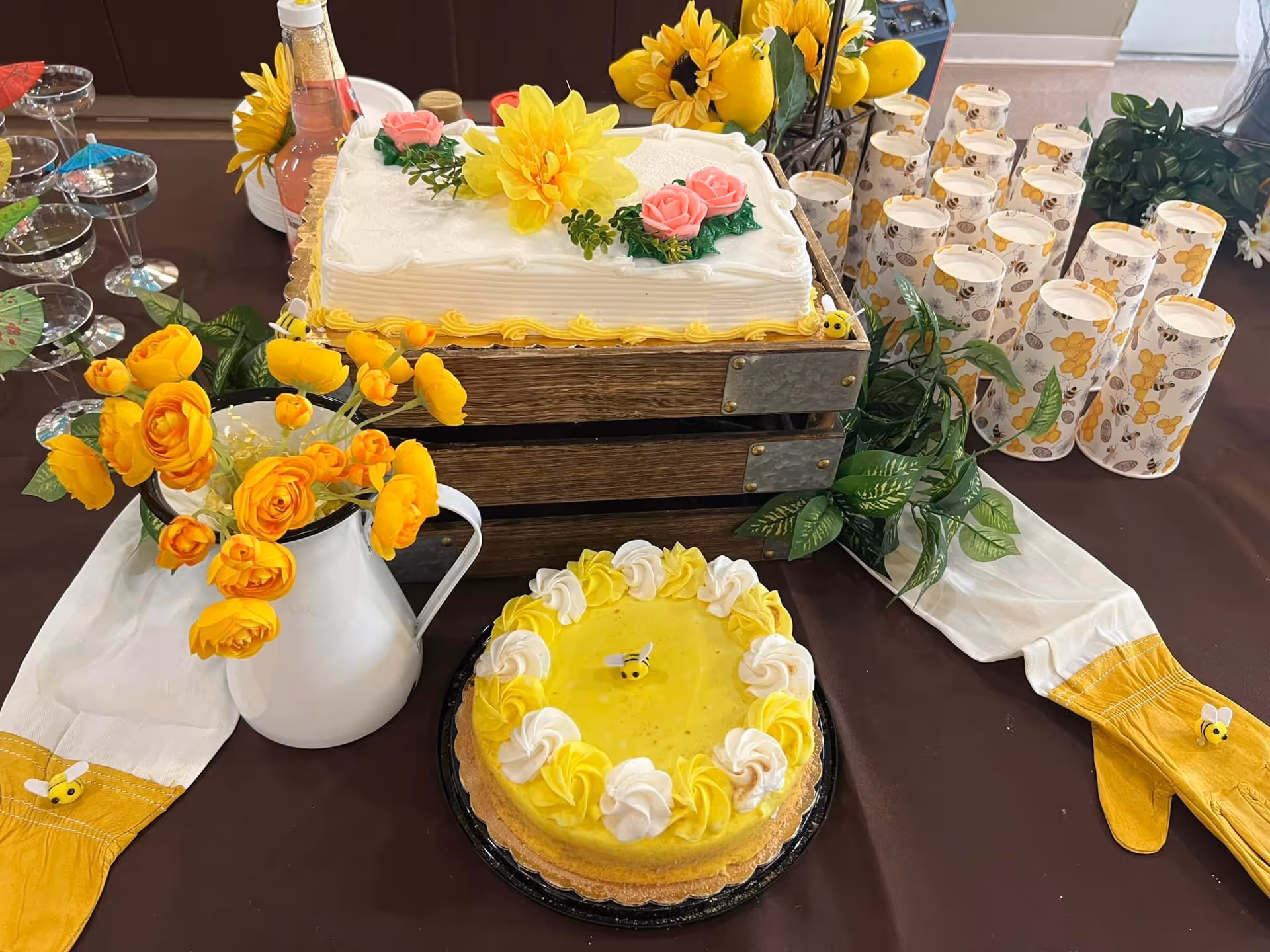 A table decorated with yellow and white themed items including two cakes, one rectangular with white frosting and yellow and pink flower decorations, and one round with yellow and white frosting and a small bee decoration. There is a white pitcher with yellow flowers, several paper cups with bee and flower designs, green leafy plants, and a pair of yellow and white gloves with small bee decorations on a brown tablecloth.