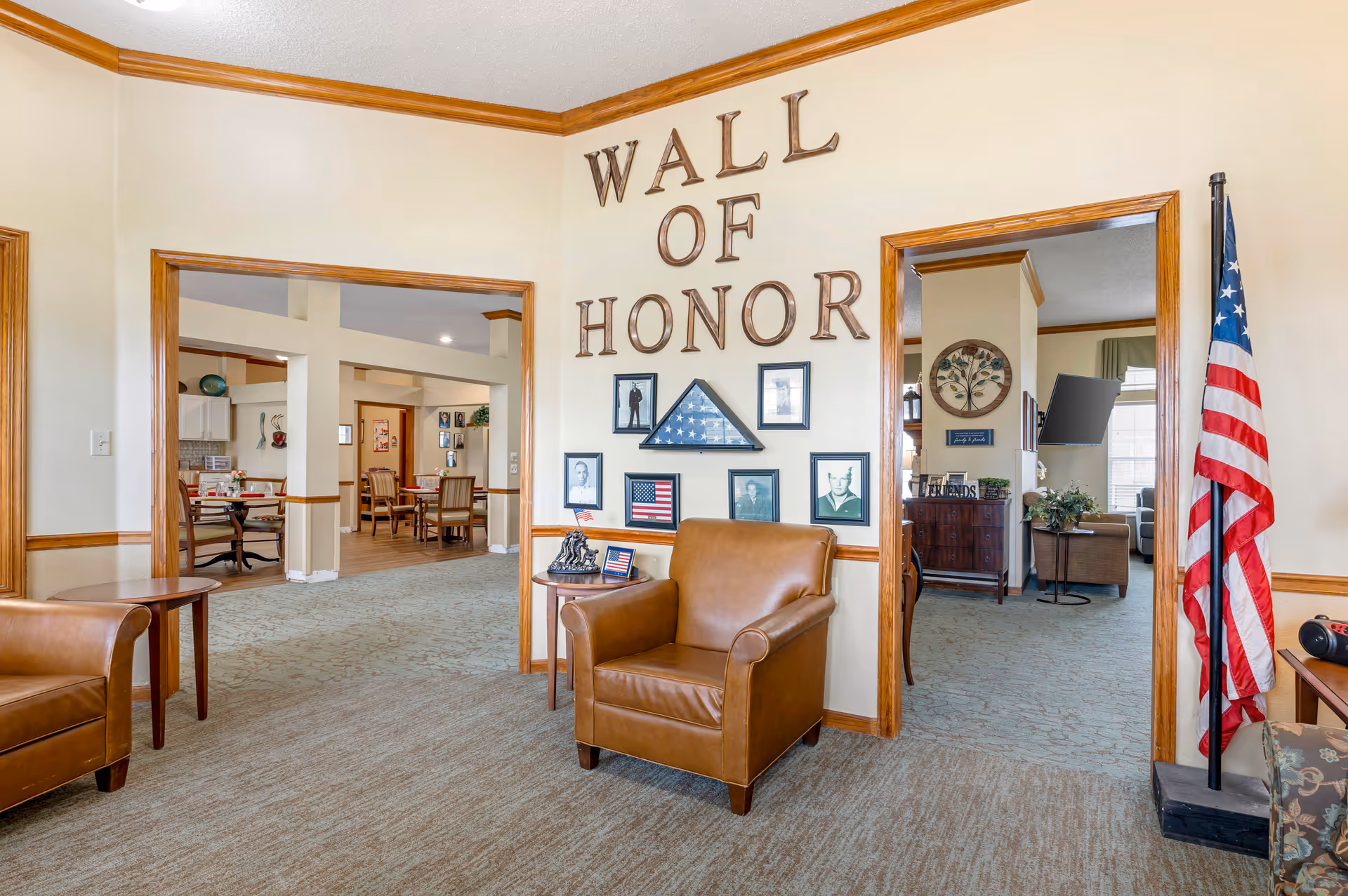 Interior view of a senior living facility featuring a 'Wall of Honor' with framed photos and a folded American flag. The room has beige walls with wooden trim, a brown leather armchair, an American flag on a stand, and doorways leading to other rooms with dining tables and chairs.