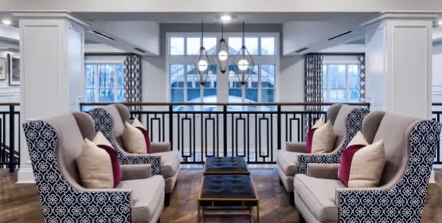 A bright and elegant common area with two pairs of patterned armchairs facing each other, each with beige and burgundy throw pillows. Between the chairs are two dark tufted ottomans. Large windows in the background let in natural light, and a modern chandelier hangs from the ceiling.