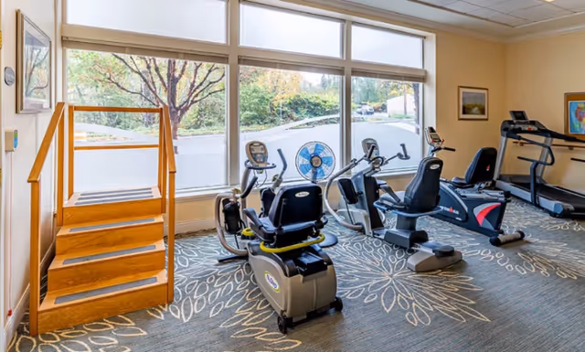 Bright senior fitness room with recumbent exercise bikes, a treadmill, a wooden stepper, and large windows overlooking trees.