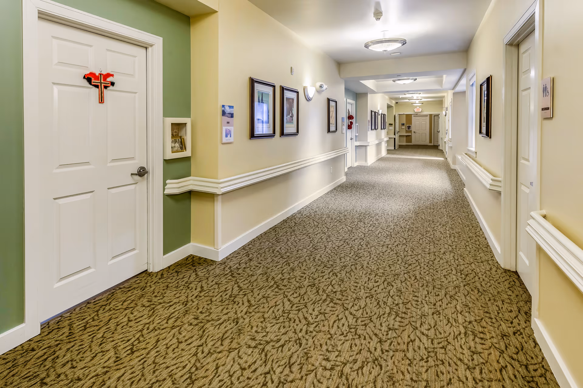 A long, well-lit hallway in a senior living facility with beige walls, patterned carpet, and white doors on both sides. The hallway is decorated with framed pictures and has handrails along the walls. One door on the left has a red and black cross decoration hanging on it.