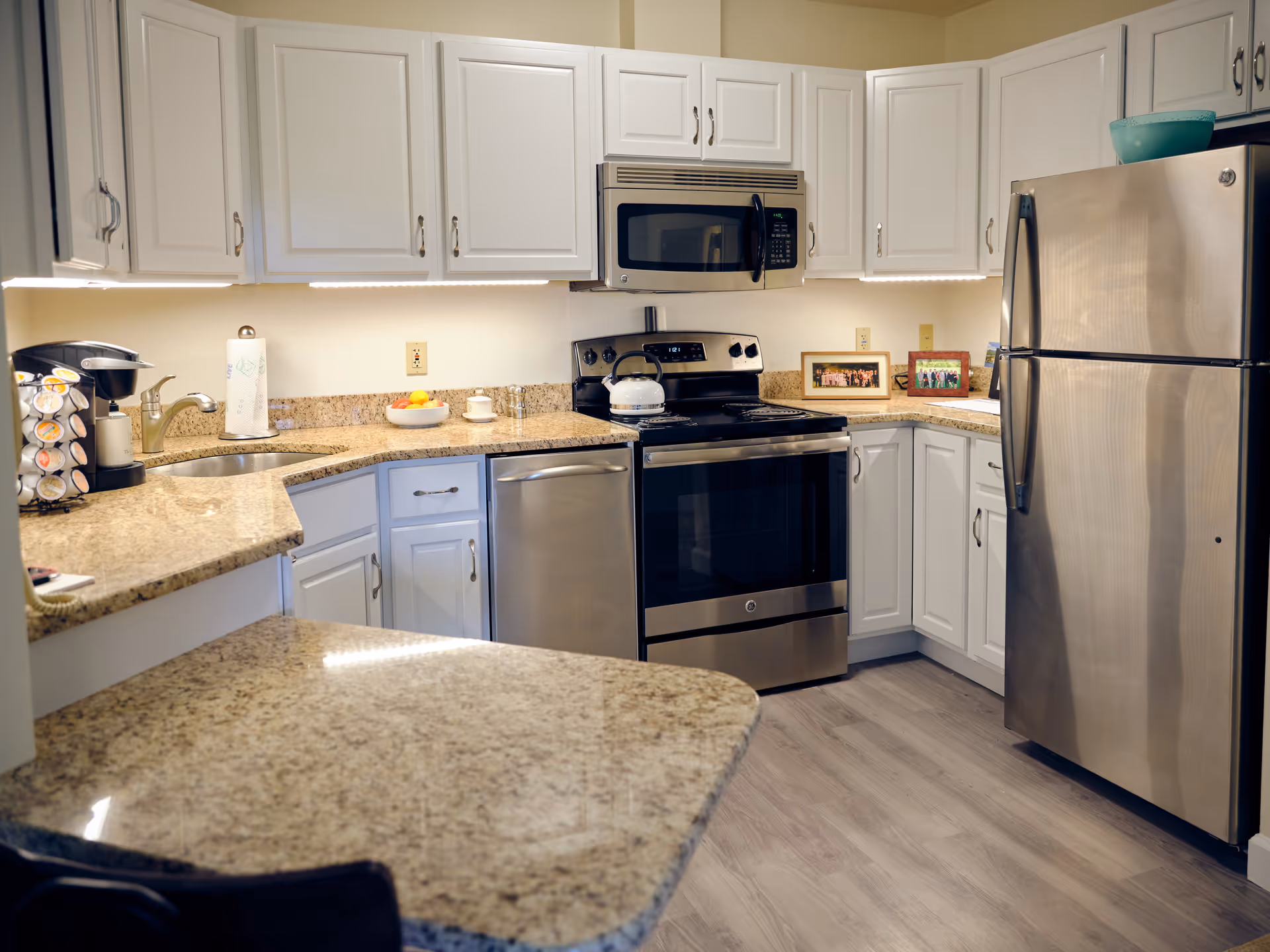 A modern kitchen with white cabinets, granite countertops, and stainless steel appliances including a refrigerator, oven, microwave, and dishwasher. There is a coffee maker on the counter, a bowl of fruit, and framed photos on the countertop near the refrigerator. The floor is light-colored wood.