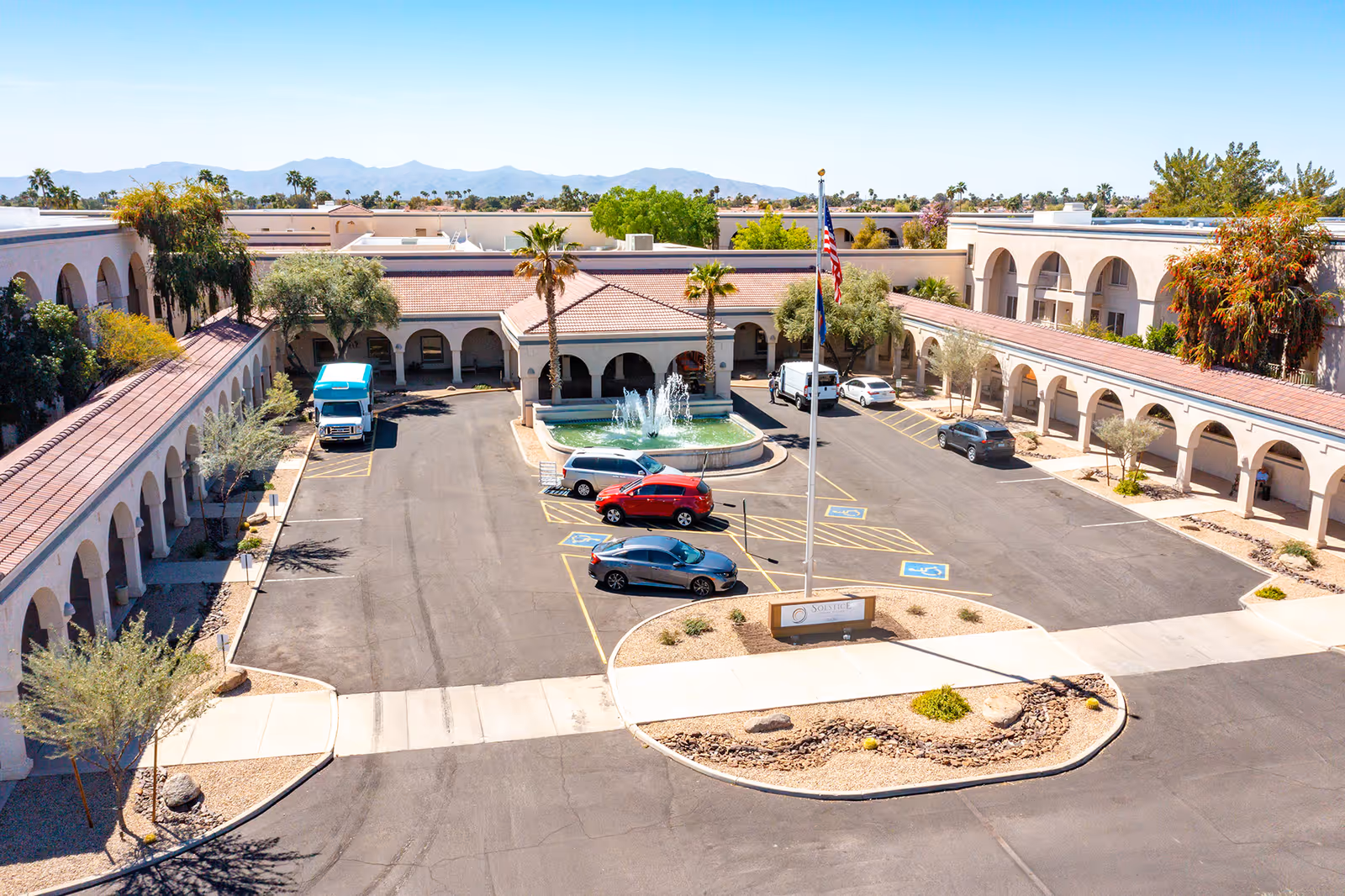 Aerial view of Solstice Senior Living at Sun City West showing a U-shaped building with arched walkways, a central fountain, several parked cars, and a flagpole with the American flag. The surrounding landscape includes desert plants and distant mountains under a clear blue sky.