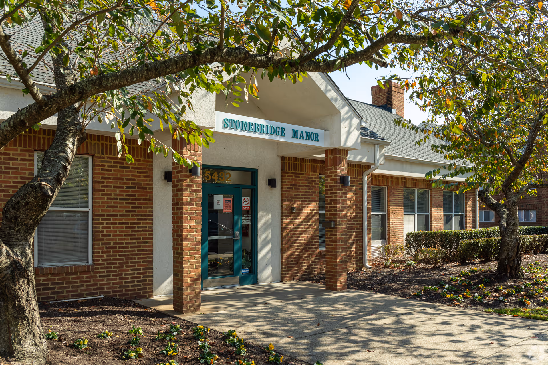 Front entrance of Stonebridge Manor with a brick facade, glass double doors, and trees and landscaping in the foreground.