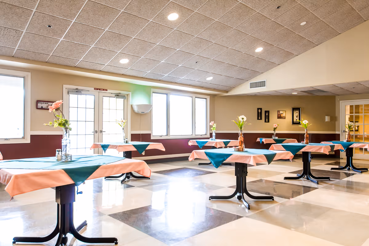 A bright dining room with multiple round tables covered with peach tablecloths and green table runners. Each table has a vase with flowers. The room has large windows, a tiled floor with a checkered pattern, and a ceiling with recessed lighting.