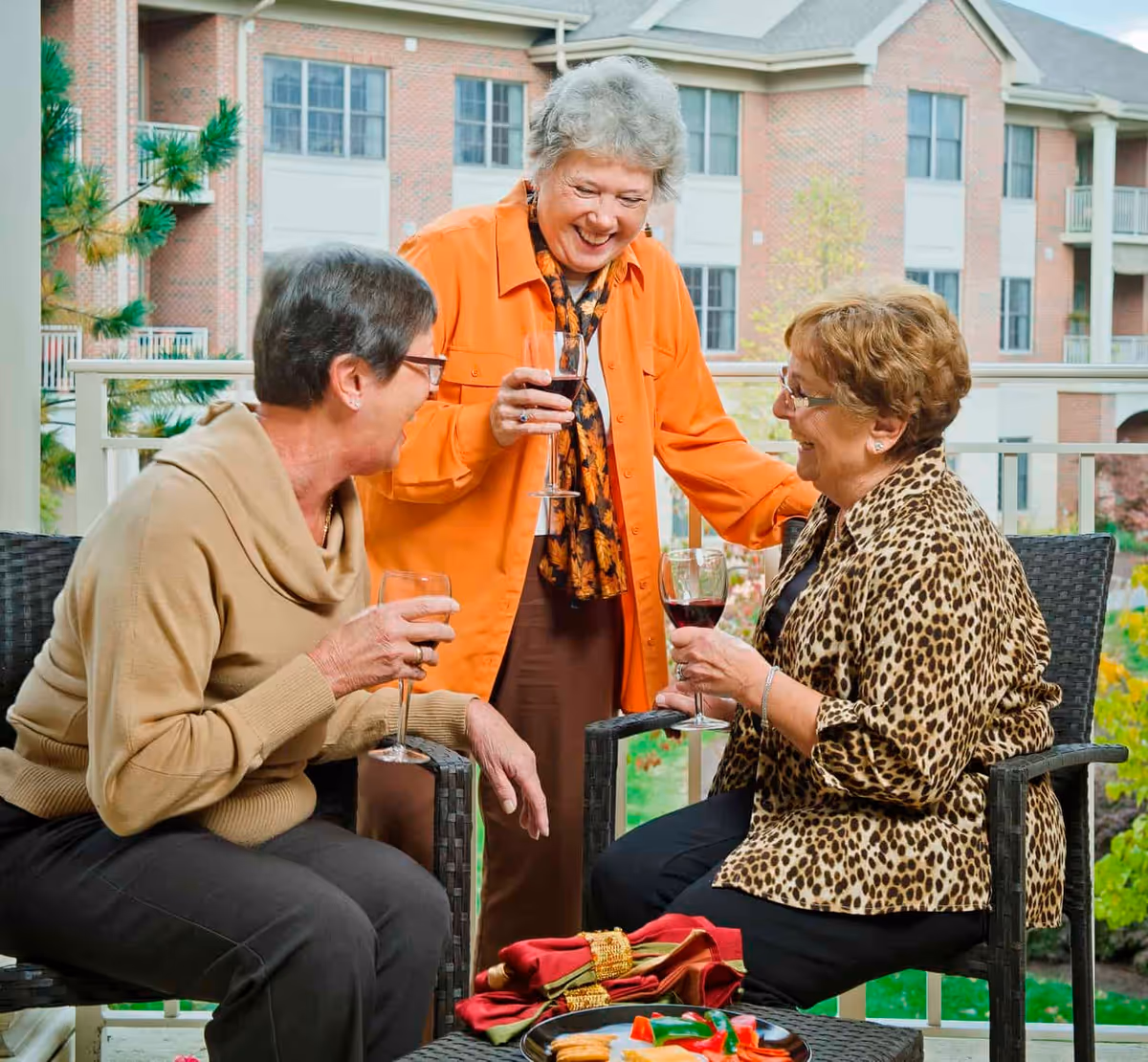 Three elderly women enjoying a social gathering on an outdoor patio. Two women are seated on wicker chairs holding glasses of red wine, while a third woman stands between them, smiling and holding a glass of wine. The background shows a brick building with multiple windows and balconies, and some greenery.