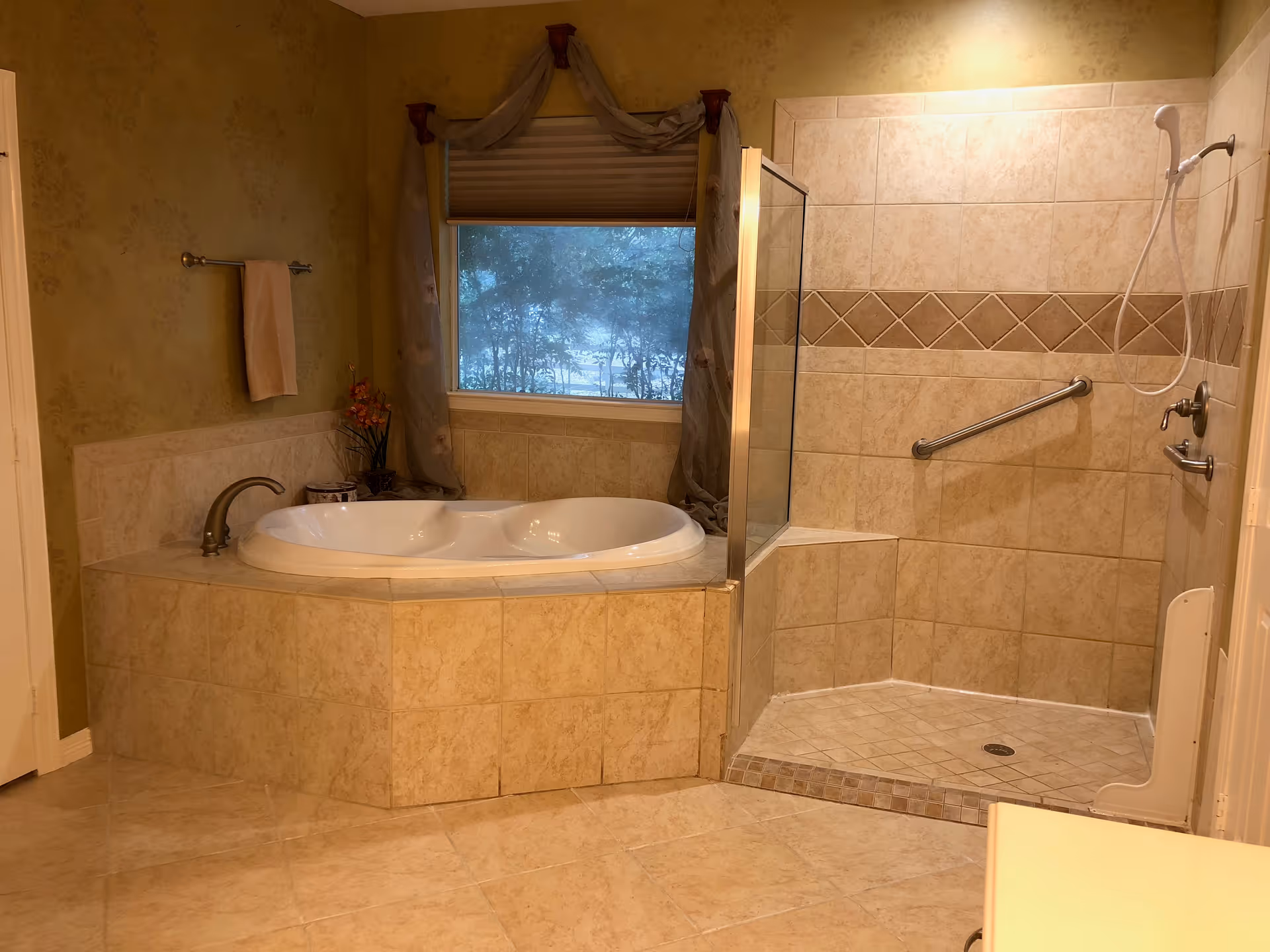 A spacious bathroom featuring a large corner bathtub with tiled surround and a separate walk-in shower with a glass partition and grab bar. The walls are decorated with a subtle floral pattern, and there is a window with a curtain above the bathtub. A towel hangs on a rack on the left wall.