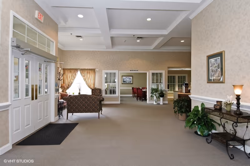 A spacious and well-lit interior common area of Whispering Pines Village featuring beige walls, carpeted floor, and a coffered ceiling with recessed lighting. There are double white entrance doors on the left, a seating area with patterned armchairs near a window with checkered curtains, and a hallway leading to a dining room with red chairs. Decorative plants and a small table with a lamp and framed artwork are on the right side.