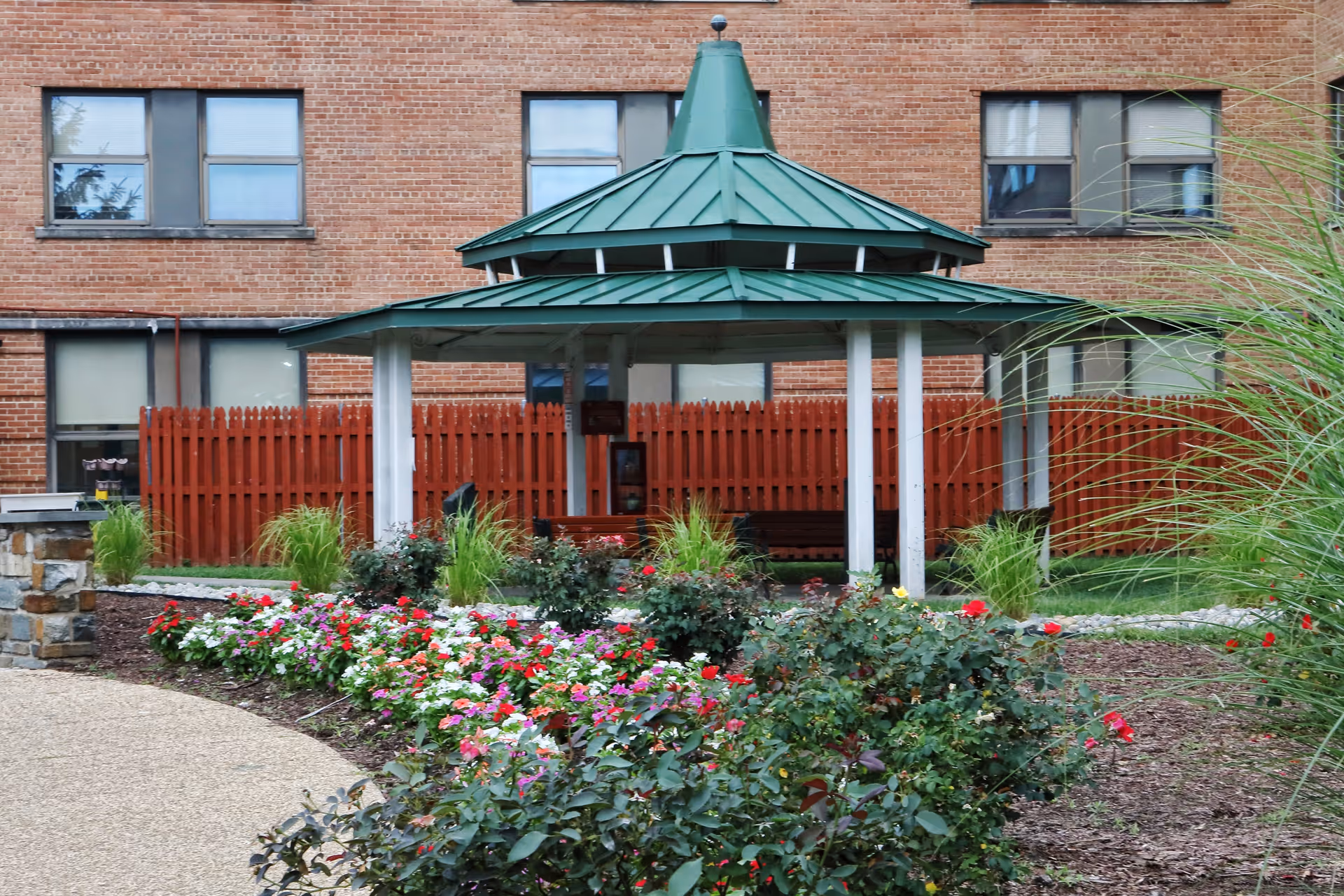 Outdoor garden area with a green-roofed gazebo surrounded by colorful flowers and plants, with a red wooden fence and a brick building in the background.