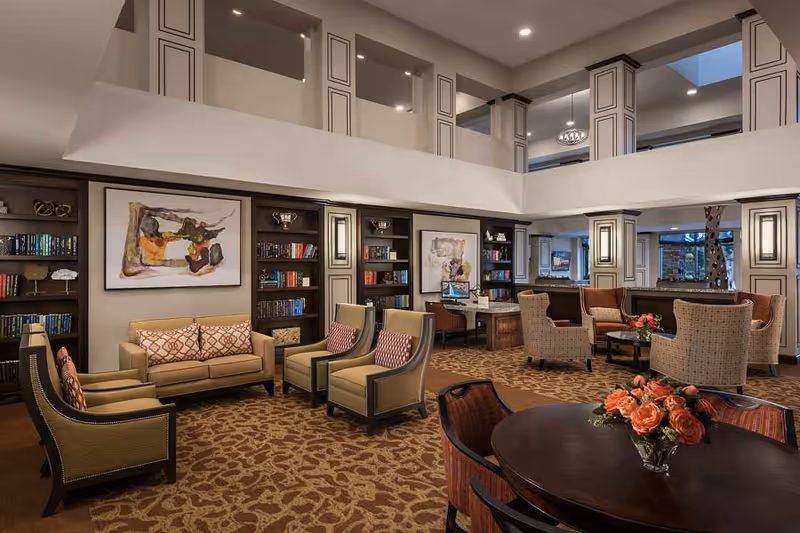 A spacious and elegant living room area in a senior living facility with high ceilings and a mezzanine level. The room features multiple seating arrangements including sofas and armchairs with patterned cushions, dark wood bookshelves filled with books and decorative items, framed abstract artwork on the walls, and a round table with a vase of orange roses in the foreground. The lighting is warm and recessed, creating a cozy and inviting atmosphere.