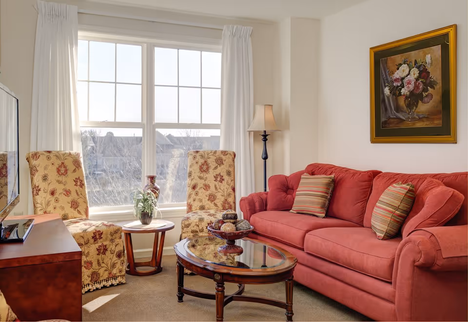 Sunny living room with a red sofa, two patterned armchairs, a glass-topped coffee table, and a large window.
