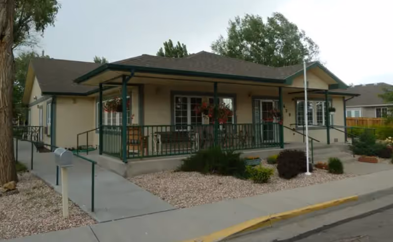 Single-story assisted living building with a covered front porch, green railing, and landscaped surroundings.