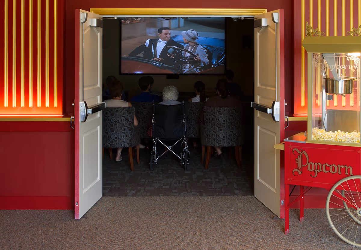 View through open double doors into a small movie theater room where several people, including one in a wheelchair, are seated watching a classic black-and-white film projected on a screen. To the right of the doors is a red popcorn machine with freshly popped popcorn inside. The walls around the doors are red with gold accents.