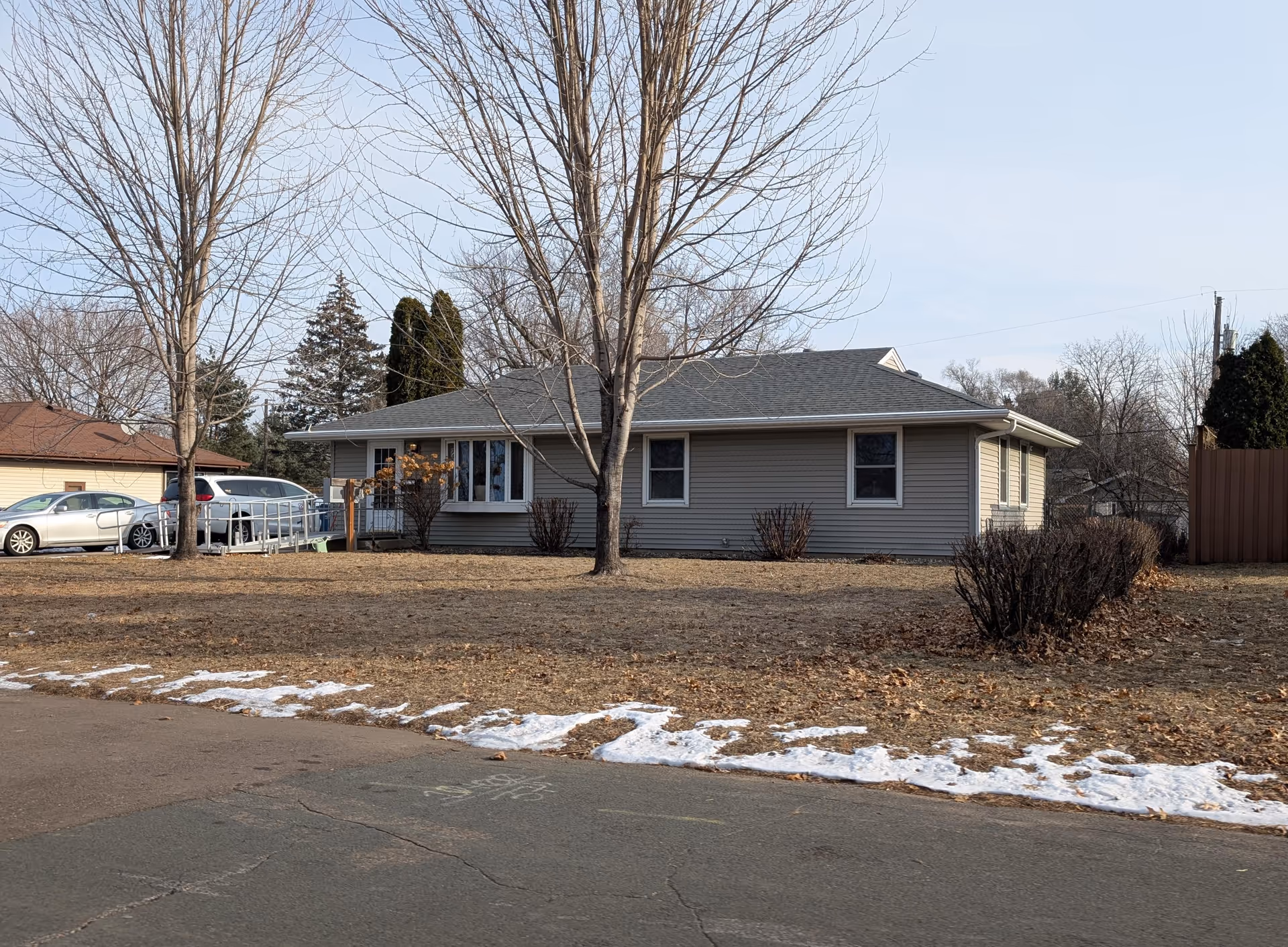 Single-story residential building with beige siding and a gray roof, surrounded by leafless trees and bushes in a yard with patches of snow on the ground. Several parked cars are visible to the left side of the building under a clear sky.