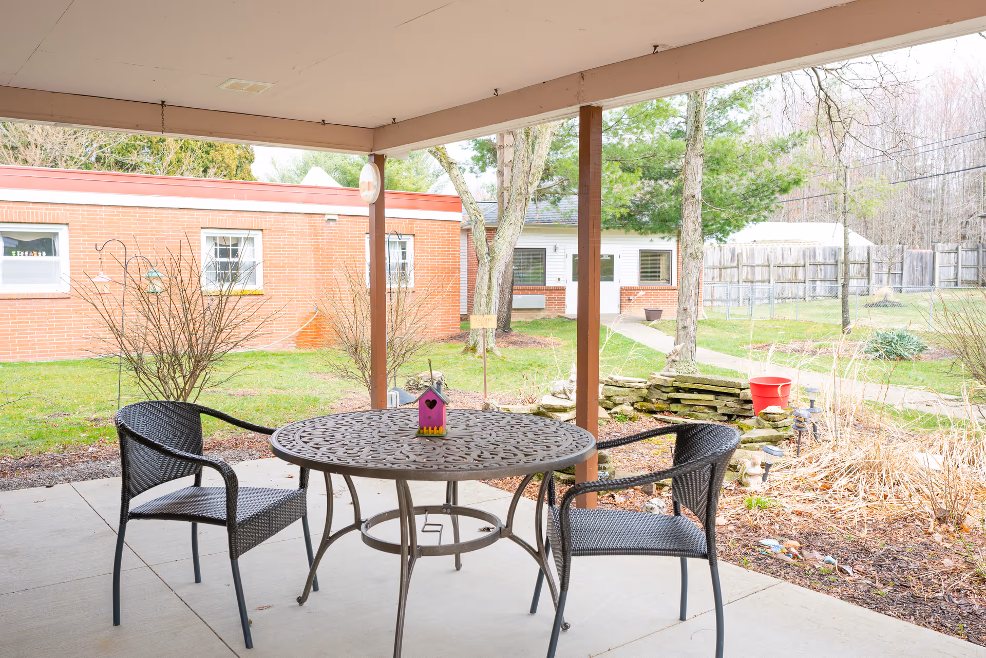 Covered outdoor patio area with a round metal table and two black wicker chairs. A small purple birdhouse is placed on the table. In the background, there is a grassy yard with trees, shrubs, a red bucket, and a brick building with windows.