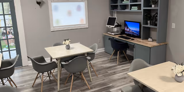 A small room with two square tables each surrounded by four gray chairs. There is a window with a frosted glass shade, a door with glass panels, and a built-in desk area with a computer and office chair. The floor is wood-patterned and the walls are painted light gray.