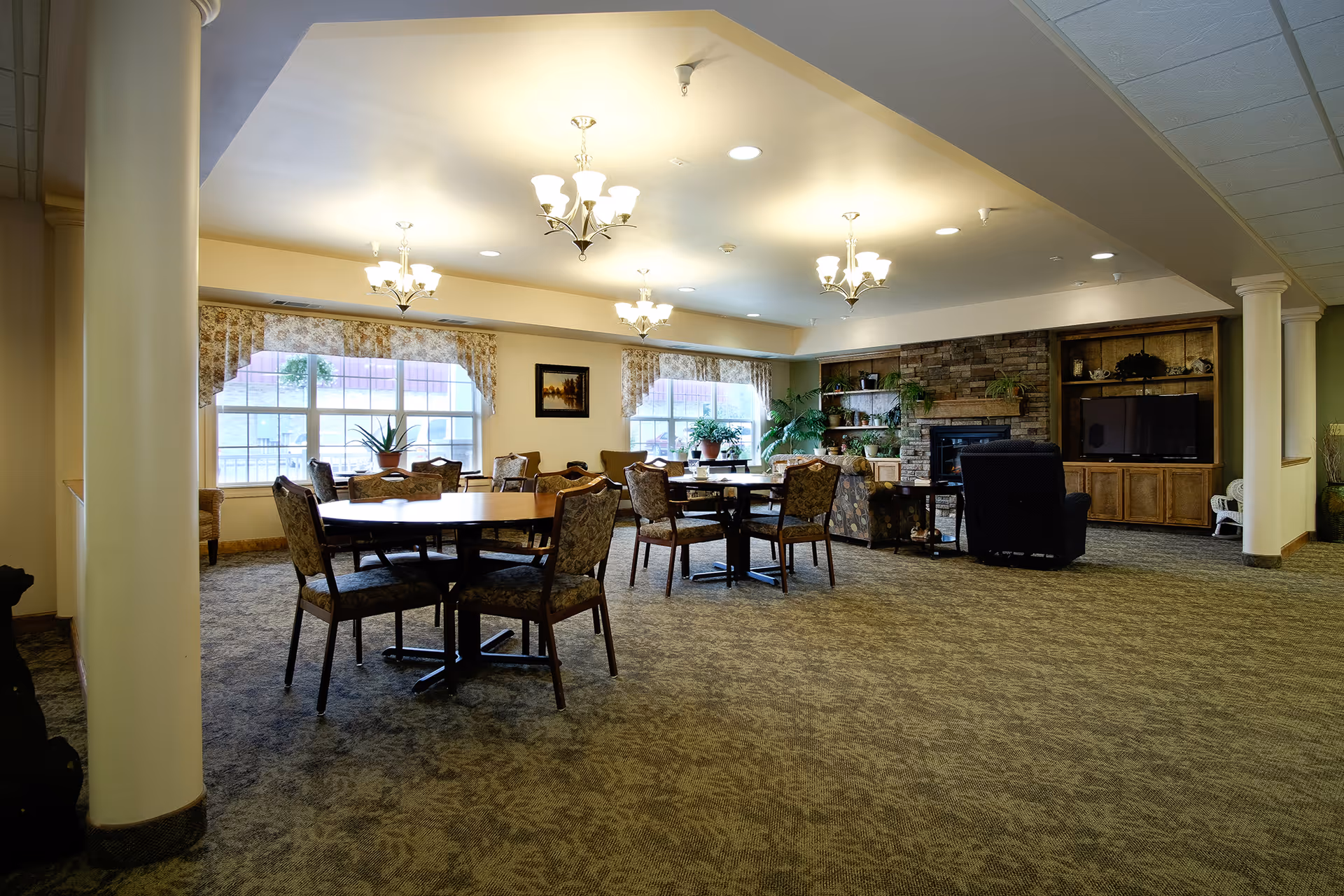 A spacious, well-lit common area in an assisted living facility featuring multiple round tables with chairs, large windows with floral curtains, a stone fireplace, a television set in a wooden cabinet, and several potted plants. The room has carpeted flooring and multiple ceiling light fixtures.