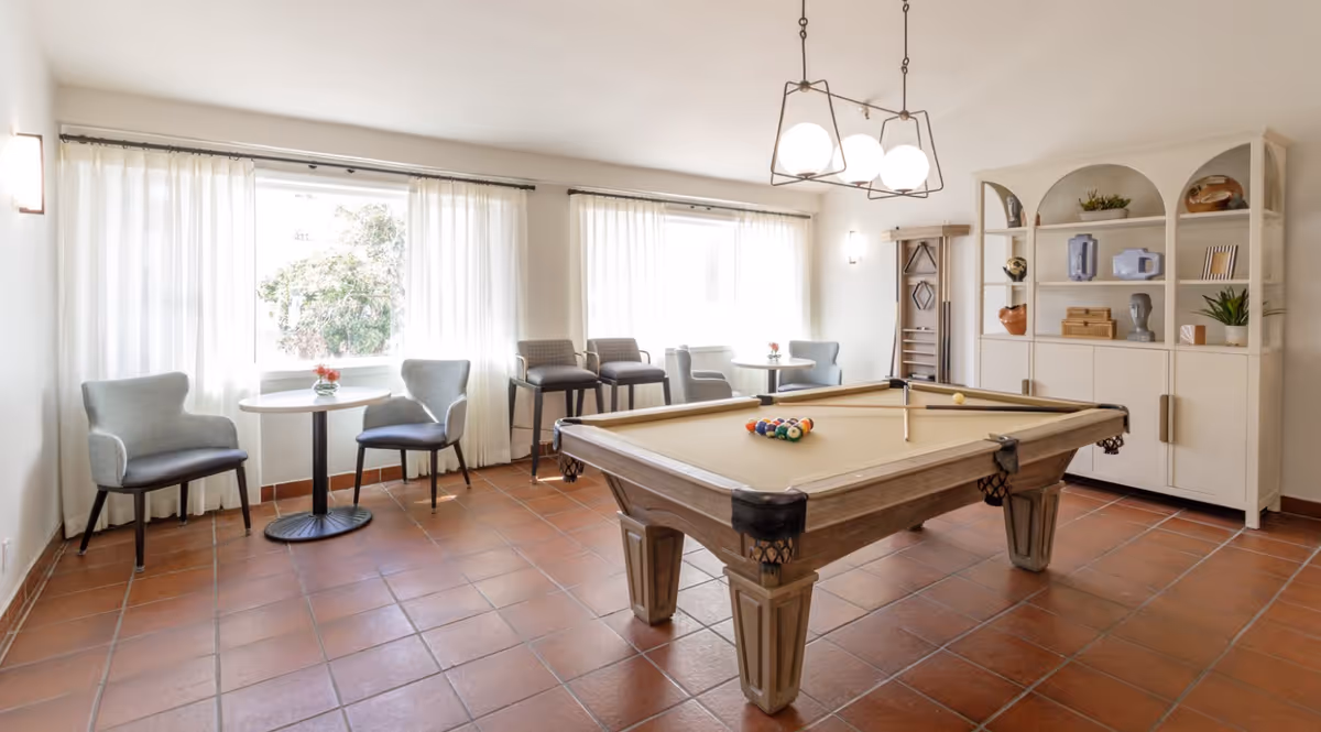 A bright room with a pool table in the center, surrounded by several chairs and small round tables near large windows with sheer white curtains. A white shelving unit with decorative items is against the wall, and a modern light fixture hangs above the pool table.