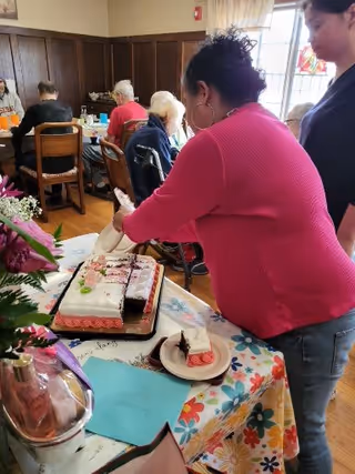 A woman in a pink sweater cuts a decorated sheet cake on a floral-covered table while other residents sit at tables in a communal dining room.