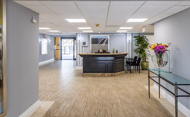 Reception area of Grand Court Lakes facility with a dark wooden front desk, a person seated behind the desk, light-colored tiled floor, gray walls, a glass table with a vase of colorful flowers on the right, and a large potted plant next to the desk. The entrance door is visible in the background.