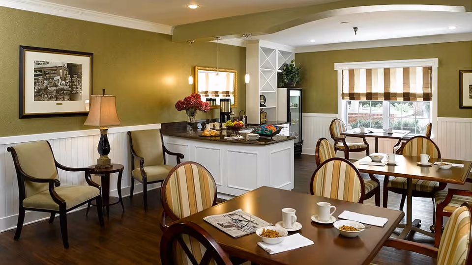 A cozy dining area in a senior living facility with several wooden tables and striped cushioned chairs. Each table has white cups, bowls with cereal, and napkins. The room features green walls with white wainscoting, framed pictures, and a window with striped blinds letting in natural light. There is a small counter area with a coffee station, flowers, and snacks, along with two armchairs and a small side table near the wall.