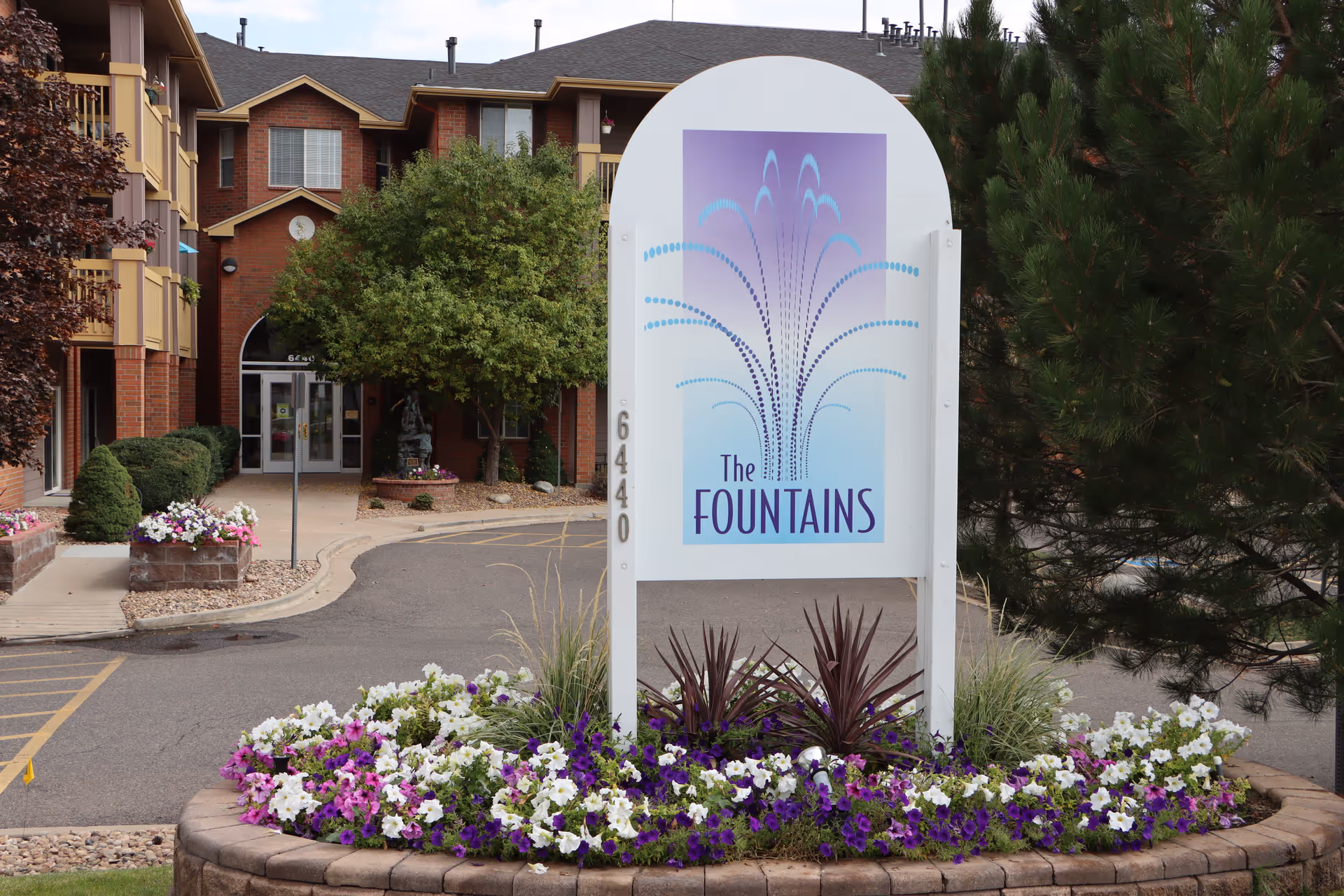 Entrance area of a senior living facility with a large sign reading 'The Fountains' surrounded by a flower bed with purple and white flowers. The building is visible in the background with brick and beige exterior, trees, and a driveway leading to the entrance.