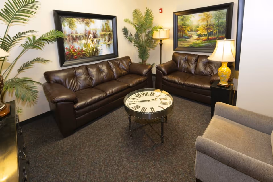Small sitting area with two brown leather sofas, an armchair, a round clock-faced coffee table, lamps, potted plants and framed landscape paintings on the walls.