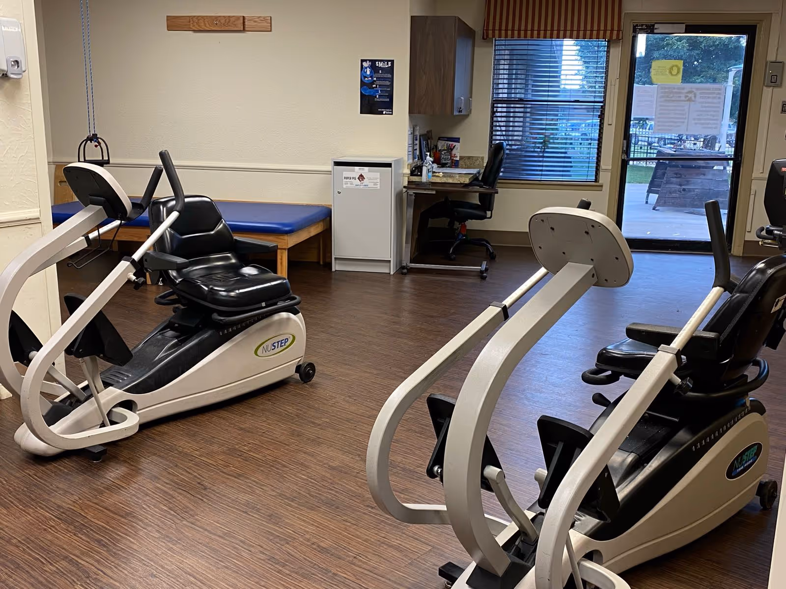 A small exercise room with two NuStep recumbent cross trainers, a blue padded bench against the wall, a desk with a chair and office supplies, and a door leading outside with windows showing an outdoor area.