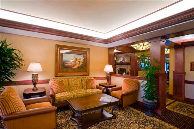 A cozy living room area in The Stafford Retirement Community featuring a patterned carpet, a wooden coffee table, a floral upholstered sofa, two striped armchairs, two table lamps on side tables, a framed painting on the wall, and a large potted plant near wooden columns.