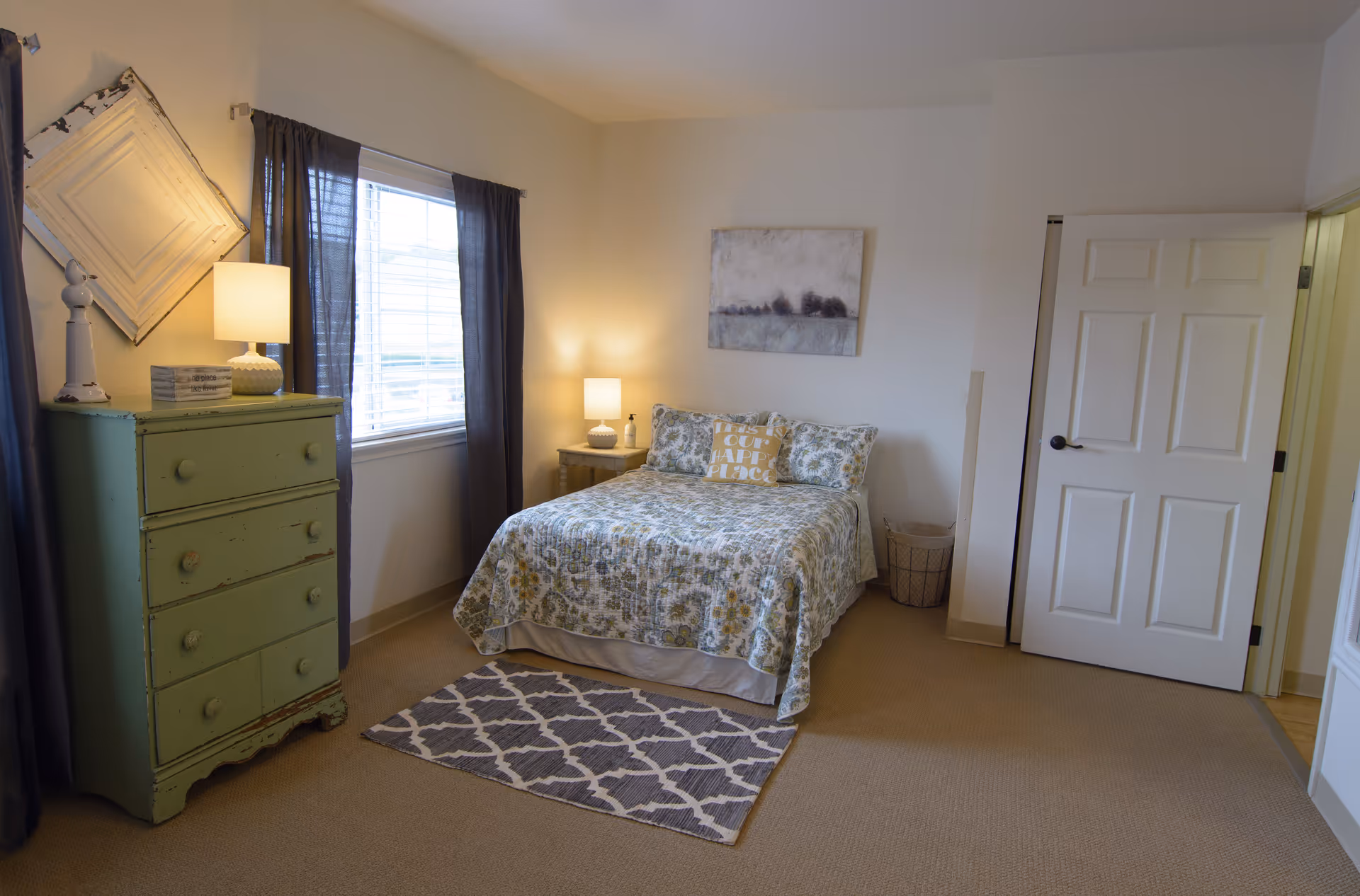 A cozy bedroom with a bed covered in a floral patterned quilt and pillows, including one with the text 'This is our happy place'. There is a green distressed dresser with a lamp and decorative items on top, a window with dark curtains, a small bedside table with a lamp, a wastebasket, a patterned rug on the carpeted floor, and a door slightly ajar leading to another room.
