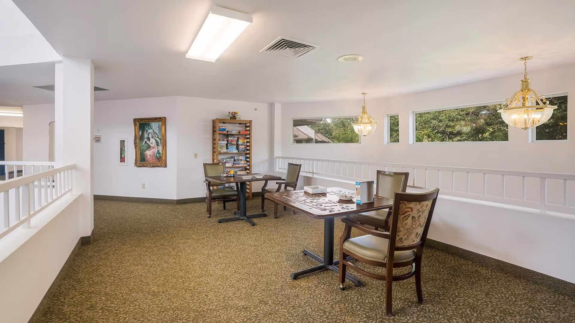 A well-lit common area with two tables and several chairs arranged around them. One table has a partially completed jigsaw puzzle and a box of puzzle pieces. Behind the tables, there is a bookshelf filled with board games. The walls are white with decorative molding, and there are three rectangular windows letting in natural light. Two hanging light fixtures with glass shades are visible. A framed painting is hung on the far wall.