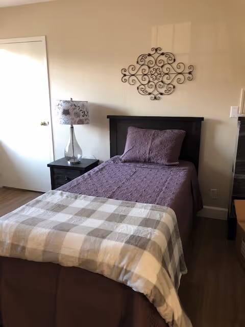 A neatly made single bed with a purple quilt and matching pillow, topped with a beige and white checkered blanket at the foot. Next to the bed is a dark wooden nightstand with a glass lamp featuring a floral lampshade. Above the bed is a decorative black wrought iron wall art. The room has light-colored walls and wooden flooring.