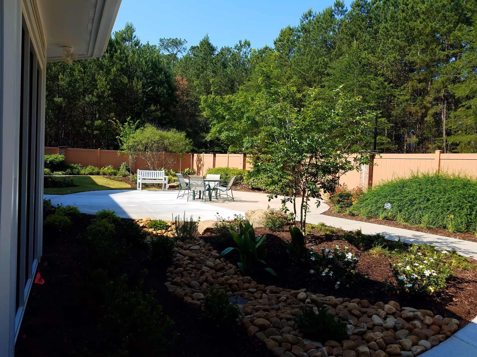 Outdoor patio area with a concrete seating space featuring a white bench and a glass table with four chairs, surrounded by landscaped garden beds with bushes, small trees, and a rock-lined dry creek bed, enclosed by a tan privacy fence and backed by tall pine trees under a clear blue sky.