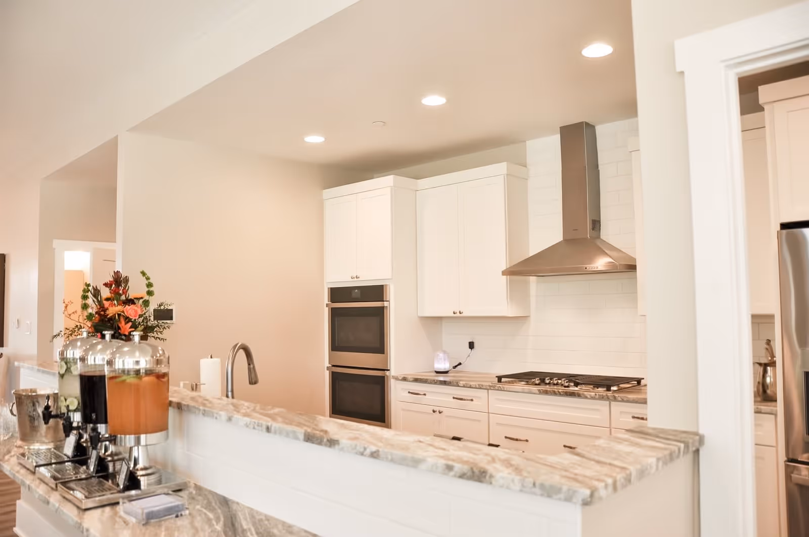 Bright modern kitchen with white cabinetry, stainless steel range hood and double wall ovens, marble countertops, and beverage dispensers on the counter.