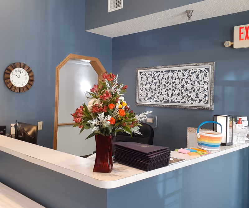 Reception desk in a blue-painted interior with a vase of flowers, stacked folders, wall clock, decorative wall art and an exit sign.
