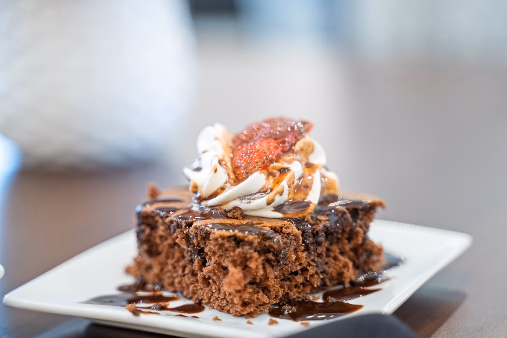 A close-up of a chocolate brownie dessert topped with whipped cream, a sliced strawberry, and drizzled with chocolate sauce on a white square plate.