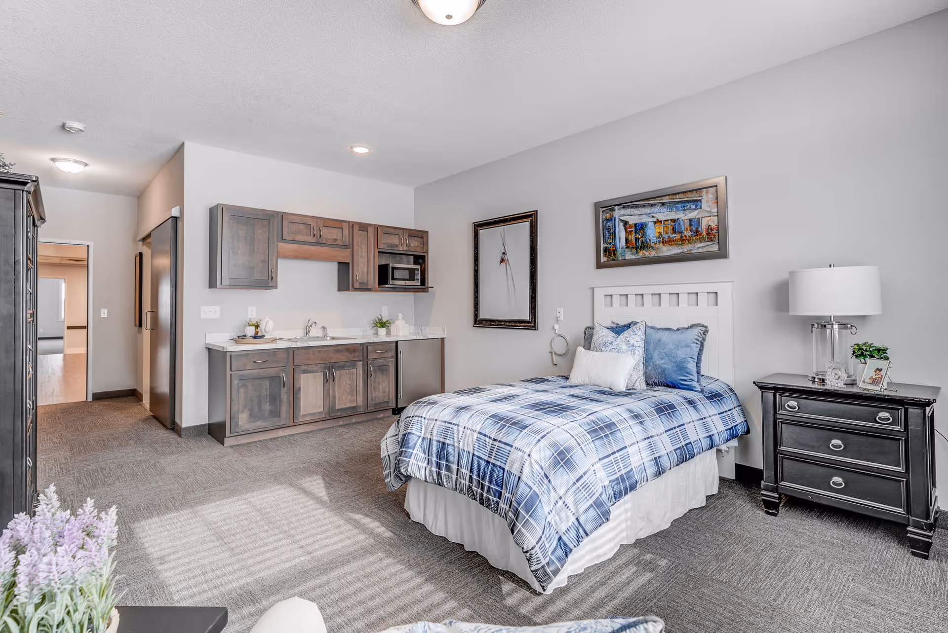 A bright and spacious senior living bedroom featuring a single bed with blue and white plaid bedding, a black nightstand with a lamp and small plant, and a kitchenette area with wooden cabinets, a sink, and a microwave. The room has light gray walls, carpeted flooring, and framed artwork on the walls.